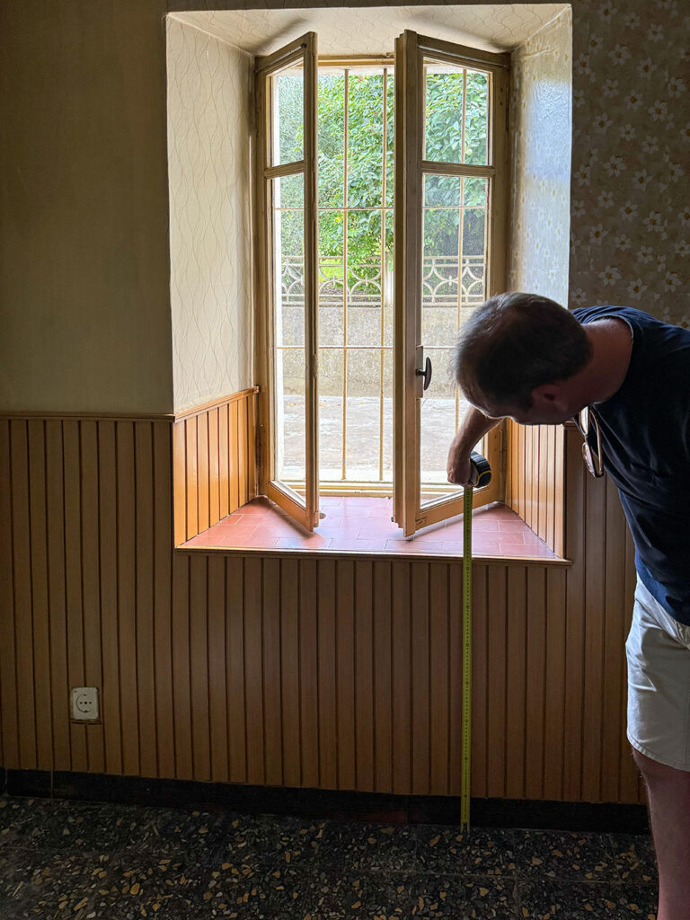 Een man meet handmatig de hoogte van de borstwering bij een raamkozijn met een klassieke rolmaat in een oude boerderij. Dit illustreert de beperkingen en foutgevoeligheid van traditioneel inmeten vergeleken met 3D-scannen.