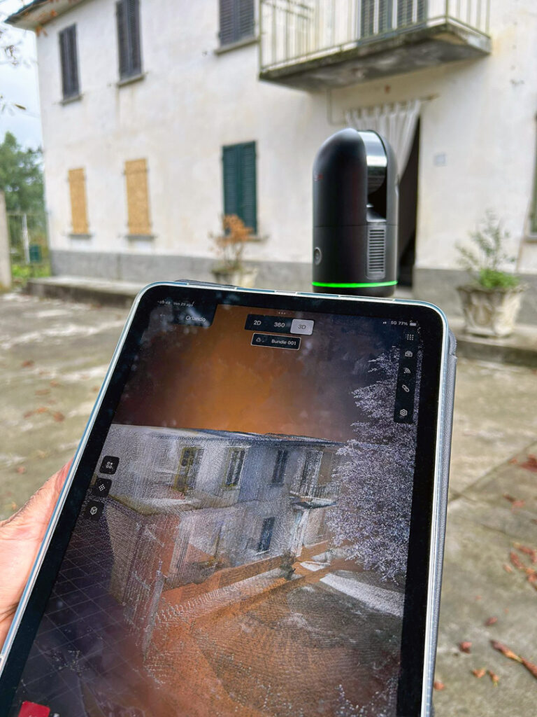 Een iPad toont real-time de 3D point cloud scan van de boerderij, terwijl op de achtergrond de Leica BLK360 scanner het exterieur inmeet. Dit demonstreert de directe controle van scandata op locatie.