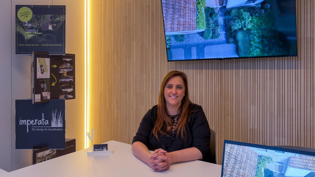 Een portretfoto van tuinontwerpster Marlies van Imperata 3D Design aan een bureau in haar studio. Op het presentatiescherm achter haar is een 3D-vogelvluchtperspectief van een tuinontwerp met oprit en beplanting te zien.