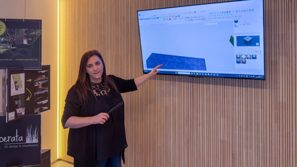Marlies van Imperata geeft een presentatie in haar studio en wijst naar een groot scherm aan de houten wand. Op de televisie is de interface van SketchUp te zien, waarmee ze het 3D-ontwerpproces live aan klanten illustreert.