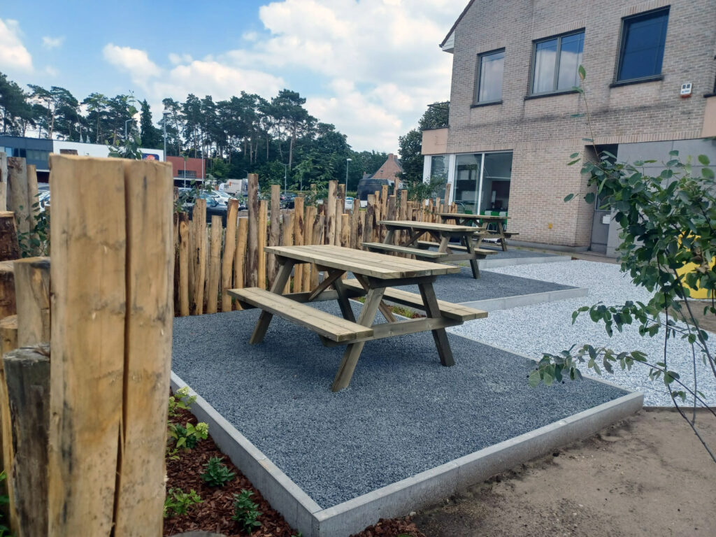 Een foto van de gerealiseerde kantoortuin. Het beeld toont de werkelijkheid die overeenkomt met het 3D-ontwerp: de houten picknicktafels, de grijze split-ondergrond en de kenmerkende afscheiding van kastanjehouten palen.
