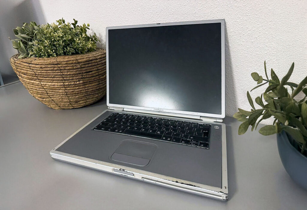A photo of a classic silver Apple PowerBook G4 laptop from 2001. The retro laptop is open on a desk next to two green houseplants, symbolizing the early hardware and workspace of Design8.