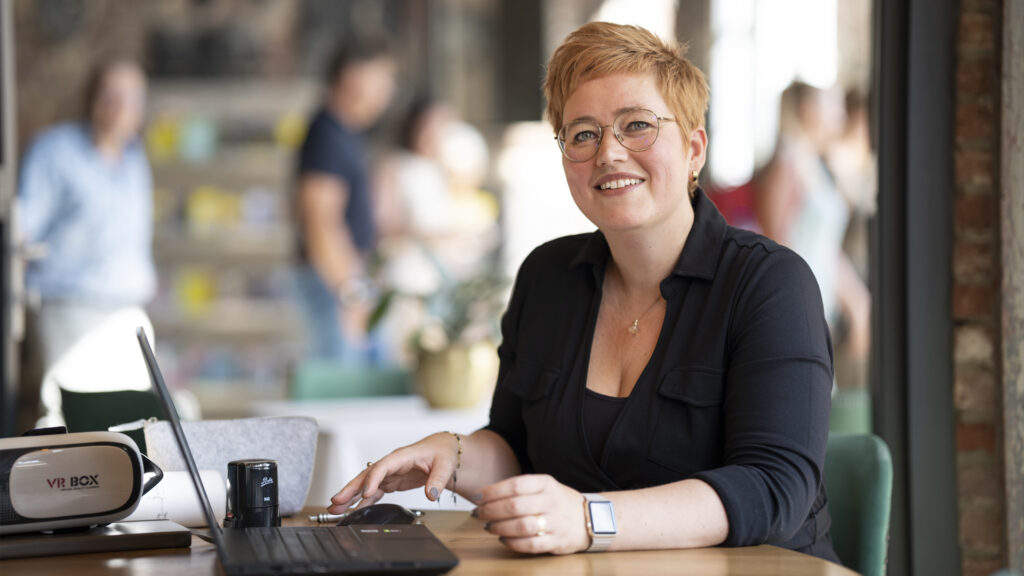 Interieurontwerper Angelique Philips zit aan een houten tafel in een sfeervolle, vervaagde ruimte met mensen op de achtergrond. Op tafel voor haar staan een geopende laptop en een 'VR BOX' Virtual Reality bril klaar, wat de moderne praktijk van interactief 3D-interieurontwerp en virtuele klantpresentaties illustreert.