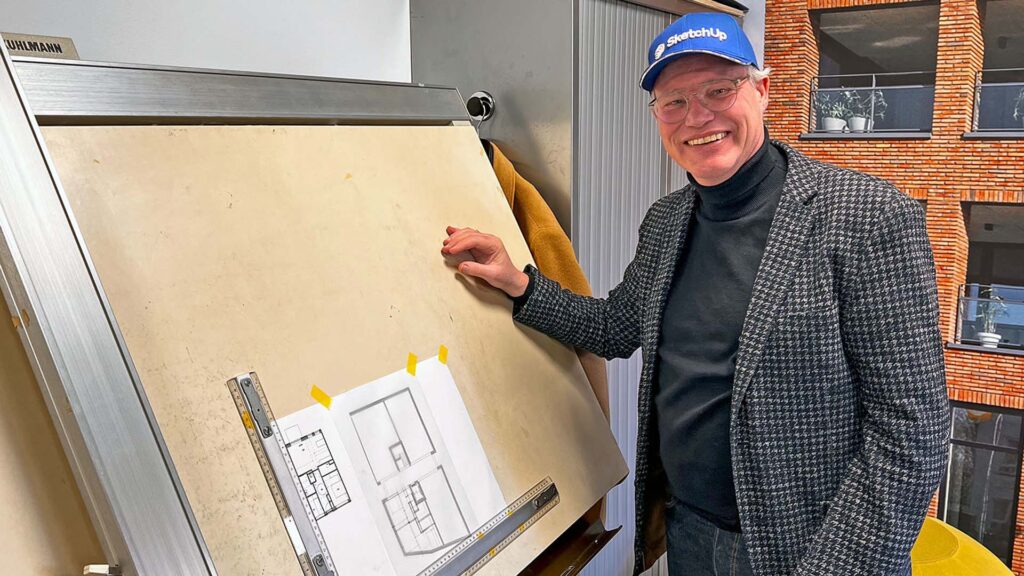 Berto van Leeuwen staat met een blauwe SketchUp-pet trots naast een klassiek houten Kuhlmann tekenbord. Op het tekenbord zijn met gele tape geprinte 2D-plattegronden bevestigd, wat prachtig de brug slaat tussen digitaal 3D-ontwerpen en de traditionele bouwkundige tekeningen voor de bouwplaats.