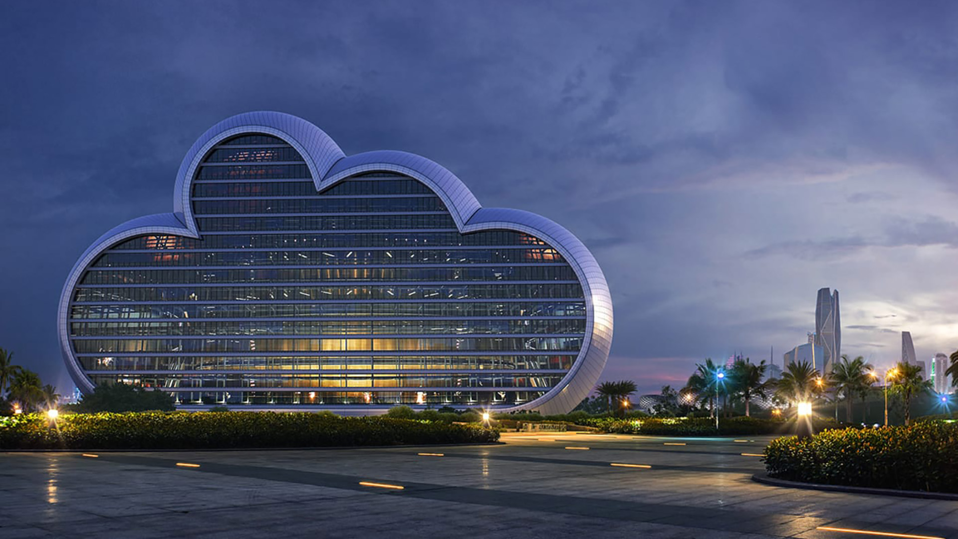 Een fotorealistische 3D-exterieurrender van een modern glazen gebouw in de vorm van een wolk, gerenderd via Chaos Cloud. Het sfeervolle beeld toont de verlichte architectuur tijdens de avondschemering, omringd door een leeg plein, palmbomen en een moderne stadsskyline op de achtergrond.