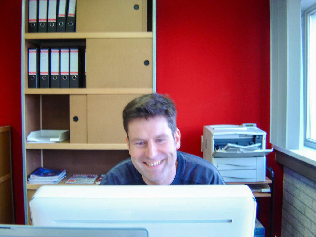 Een archieffoto van een lachende Ben van Steenwijk, achter een witte iMac op het vroege kantoor van Design8. Op de achtergrond is een kenmerkende rode kantoormuur met ordners zichtbaar.