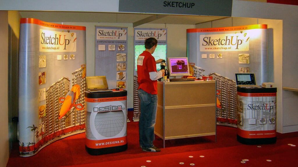 An overview photo of an early Design8 exhibition stand promoting SketchUp, featuring striking curved banners. Robert, in a red polo shirt, is neatly setting everything up on our stand at the Bouw & ICT Beurs 2006.