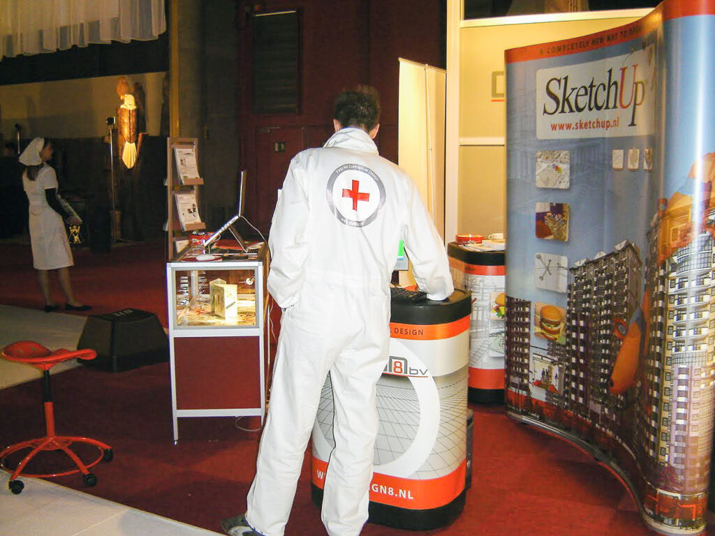 Een Design8 medewerker in een witte overall met een rood kruis en de tekst 'First Aid Conceptual Design' op een vroege SketchUp beursstand. Een creatieve marketingactie uit de beginjaren van de software distributeur.