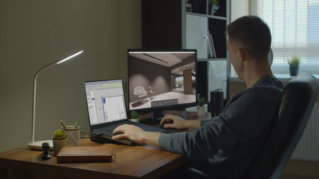 A 3D artist works at a wooden desk with a laptop and an external monitor. The laptop displays the interface of a technical CAD drawing, while the large monitor shows the realistic 3D interior render of that same space. This image illustrates a professional's workflow, fitting the choice between learning to render yourself or outsourcing it.