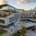 Een indrukwekkende, fotorealistische architectuurvisualisatie gegenereerd met Chaos Veras. Het beeld toont een spectaculair, gelaagd modern gebouw met grote glaspartijen, groene terrassen, wandelende mensen en een stadsskyline in de achtergrond tijdens het gouden uur.