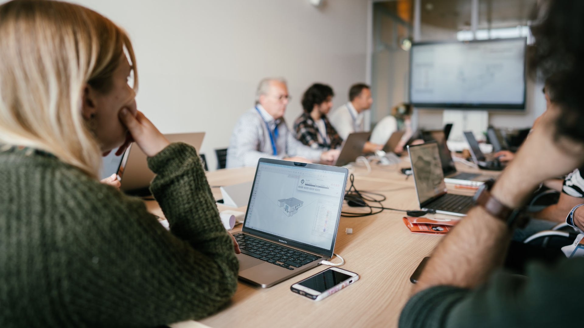 Een sfeervolle overzichtsfoto van een klassikale 3D-software training in een lichte, moderne ruimte. Op de voorgrond werkt een cursist op een laptop aan een 3D-model van een gebouw in SketchUp. Op de achtergrond volgen andere deelnemers geconcentreerd de workshop, kijkend naar hun eigen schermen en een centrale presentatiemonitor.