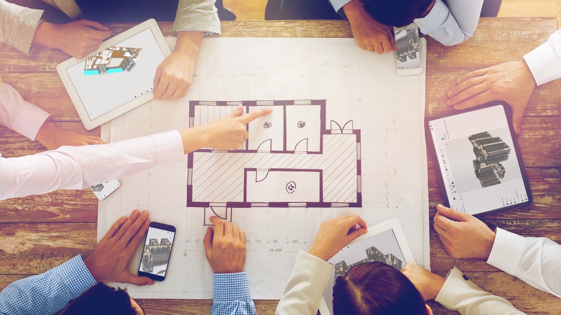 Een bovenaanzicht van een bouwteam dat samenwerkt rond een houten kantoortafel. Centraal ligt een traditionele 2D-papieren plattegrond waarnaar gewezen wordt, terwijl meerdere teamleden op hun tablets en smartphones tegelijkertijd het bijbehorende 3D-model in Trimble Connect bekijken.