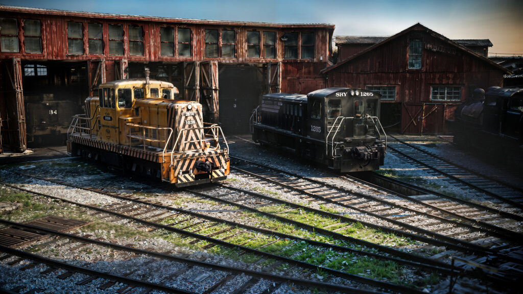 A photorealistic render of two old diesel locomotives in a weathered train yard. The image demonstrates the unprecedented 'relight' capabilities of V-Ray 7 on scanned, volumetric Gaussian Splats environments.