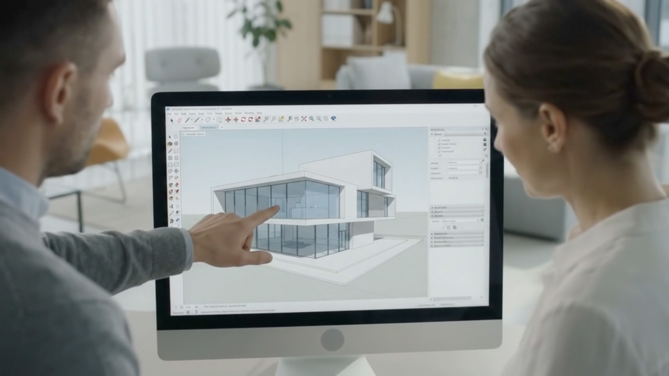 A male expert and a female trainee look at a large computer screen together in a modern office. He guides her through a complex 3D model of a contemporary villa in the SketchUp interface, illustrating a one-to-one coaching session.