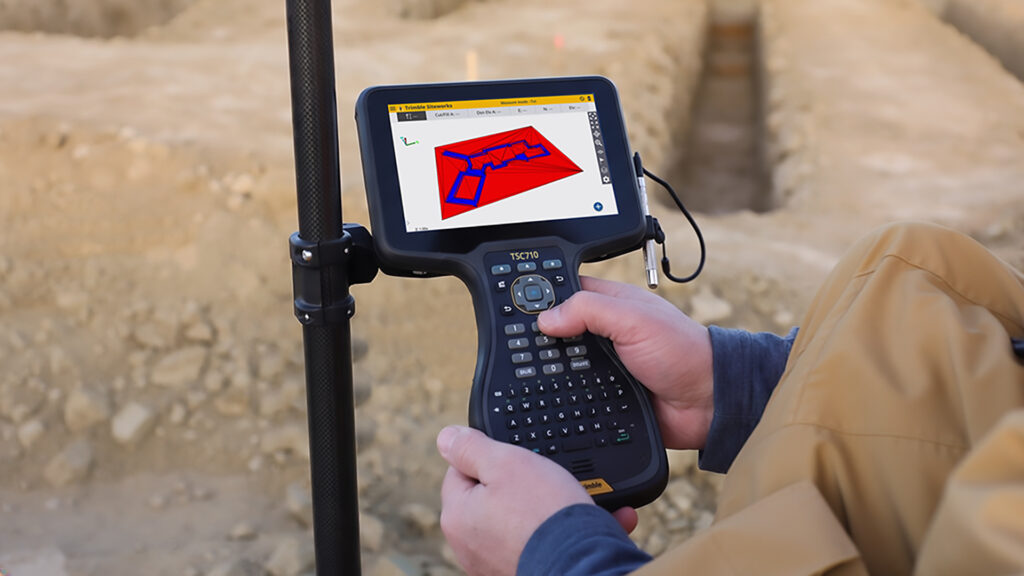 Een close-up van een persoon in werkkleding die op een zanderige bouwplaats een Trimble TSC7 veldboek bedient. Het scherm toont een helder, digitaal uitzetplan met blauwe en rode markeringen voor sleuven en funderingen.