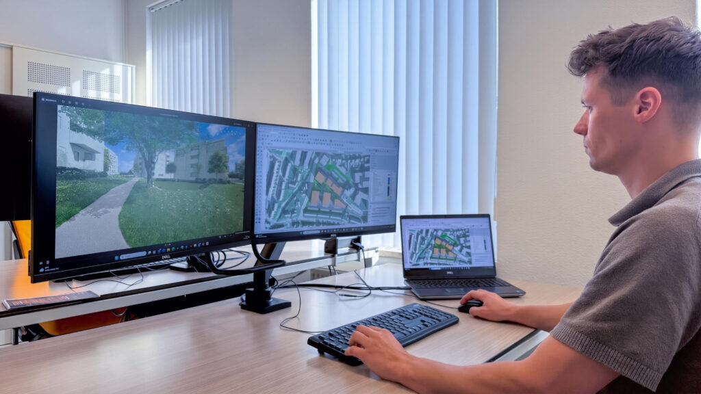 Een stedenbouwkundig ontwerper van Buro Waalbrug werkt geconcentreerd achter zijn bureau met twee grote monitoren en een laptop. Op de schermen zijn gedetailleerde 3D-modellen en plattegronden van een stedelijke omgeving te zien, ontworpen in SketchUp.