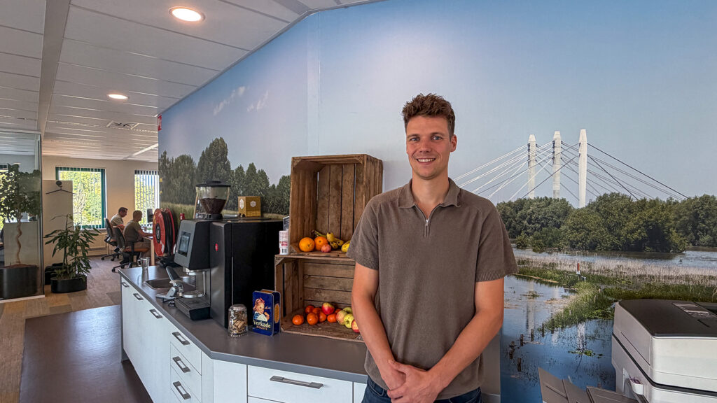 De stedenbouwkundig ontwerper poseert lachend in de kantine van Buro Waalbrug, bij het koffiezetapparaat en houten kistjes gevuld met vers fruit. Op de achtergrond bedekt een grote fotoprint van de Waalbrug in een groen rivierlandschap de muur.